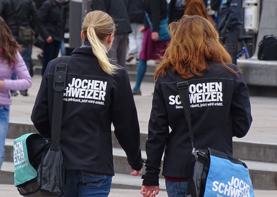 Zwei Studentinnen bei einem Promotion-Job mit schwarzen Jacken von Jochen Schweizer auf einem Platz mit vielen Menschen.