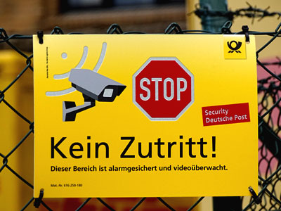 Das Foto zeigt ein gelbes Schild von der Deutschen Post mit einem Stopzeichen, einer Kamera und den Worten: Kein Zutritt! Das Bild symbolisiert die vertraulichen Informationen von Unternehmen, mit denen ein Wirtschaftsdetektiv in seinem Beruf konfrontiert wird.