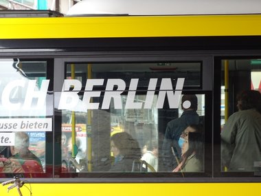 Ein Berlinschriftzug in weiß auf einer Busfensterscheibe.Im Bus sitzen mehrere Personen.