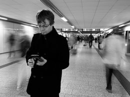 Ein Student schaut auf sein Handy auf einem schwarz weiß Bild in einem Bahnhofsgang mit verschwommenen Menschen um ihn herum und einer Anzeigentafel in der Ferne.