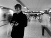 Ein Student schaut auf sein Handy auf einem schwarz weiß Bild in einem Bahnhofsgang mit verschwommenen Menschen um ihn herum und einer Anzeigentafel in der Ferne.
