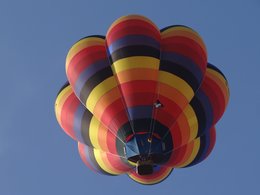  Das Bild zeigt einen Heißluftballon mit knallig bunten Streifen in gelb, orange, rot, violett und schwarz vor einem tief blauen Himmel . Darunter sieht man den Korb hängen und an der Seite eine Piratenflagge.