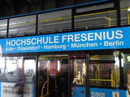 Ein blauer Banner auf einem Doppeldeckerbus mit weißer Schrift: Hochschule Fresenius- Köln, Düsseldorf, Hamburg, München und Berlin.,