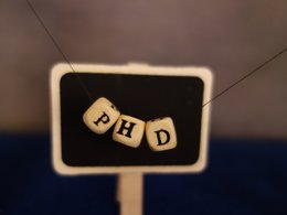 Drei Holzwürfel mit den Buchstaben PHD vor einer kleinen Schiefertafel und verschwommenem Hintergrund.