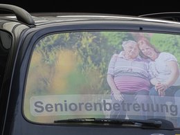 Ein Bild auf der Heckscheibe des PKW einer Seniorenbetreuung. Es zeigt eine ältere Dame lachend und eng beisamen mit ihrer Altenpflegerin draufen im Grünen sitzend.