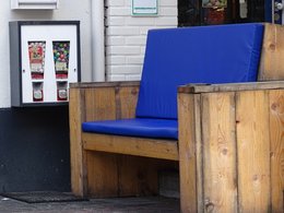 Eine Sitzbank aus Holz mit einer blauen Plastikauflage und einem Süßigkeitenautomat im Hintergrund.