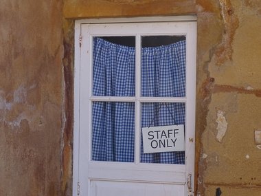Ein weißes Fenster mit blau karierter Gardiene und dem hinter der Scheibe schräg hängendem Schild: Staff only.