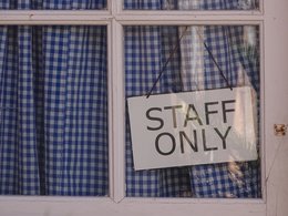 Ein weißes Fenster mit blau karierter Gardiene und dem hinter der Scheibe schräg hängendem Schild: Staff only.