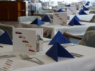 Mehrere gedeckte Tische in einem Restaurant mit blauen Servietten und einer Weinkarte in der Mitte.