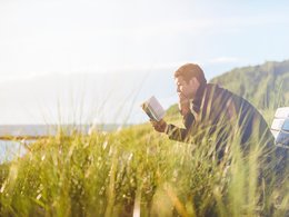 Ein Mann sitzt auf einer Bank mit Gras umgeben am Meer und liest in einem Buch.