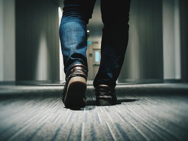 Eine Person mit braunen Schuhen und Jeans geht einen Flur entlang.