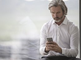 Ein Mann mit grauen Haaren, Bart und weißem Hemd sitzt am Fenster und hat ein Handy in der Hand.