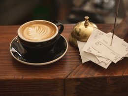 Eine Tasse Kaffee mit einem Milchschaumblatt, einer Klingel und Rechnungszettel.