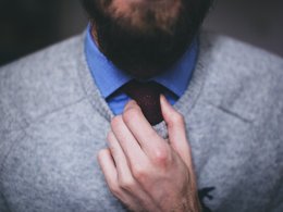 Eine Hand an einer Kravatte von einem Mann im grauen Pullover mit Bart.