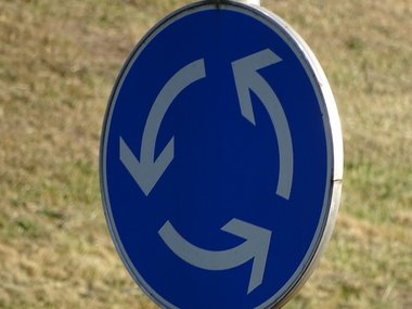 Ein blaues Kreisverkehr-Zeichen mit drei weissen Pfeilen vor einer Rasenfläche.