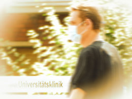 Ein Mann, der einen Mundschutz trägt mit einem Schild der Universitätsklinik im Hintergrund.