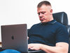 Das Bild zeigt einen Mann im Polohemd der im Büro an einem Macbook arbeitet.