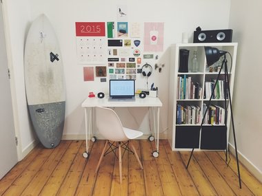 Ein Schreibtisch mit Computer mit Wandbildern, Bücherregal und Wellenreitbrett.