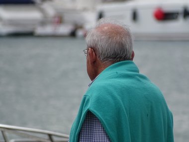 Ein Senior mit Brille, einem grauen Haarkranz und einem grünen Pullover über den Schultern.