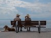 Ein Seniorenpaar mit Strohhut sitzt auf einer Bank am Meer und neben ihnen liegt ein Hund.