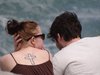 Ein Paar sitzt am Strand und sie hat im Nacken ein Kreuz tätowiert und trägt ein braunes Top und eine Sonnenbrille.