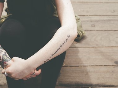Ein Arm mit einer Schrifttätowierung von einer Frau mit Ringen in schwarzer Kleidung hockend auf einem Holzfußboden.