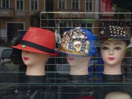 Drei Puppenköpfe mit Hüten in rot, blau und schwarz zum Teil mit goldenen Ansteckern in einem Schaufenster.