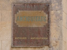Ein altes, verrostetes Schild mit der spanischen und deutschen Aufschrift für Antiquitäten hängt an einer Mauer.