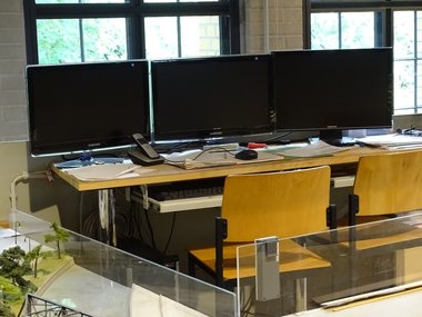 Zwei Holzstühle stehen vor einem Schreibtisch mit drei großen Monitoren und Papiermaterial und einem Telefon.