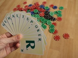 Eine Hand hält gefächerte Karten mit Buchstaben, die das Wort Gehaltspoker ergeben und im Hintergrund liegen verschwommene Pokerchips auf dem Tisch.