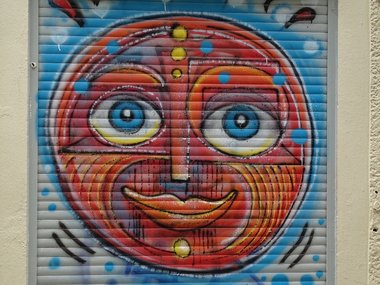 Ein rundes gemaltes Gesicht in bunten Farben auf einer geschlossenen Ja­lou­sie.