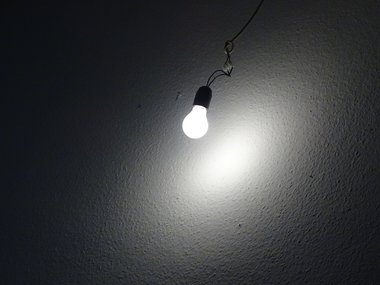Weiß leuchtende  Glühnbirne in einer Fassung an Lüsterklemme und Kabeln, die einen nach außen hin schwächer werden Lichtkreis an eine weiße Wand wirft.