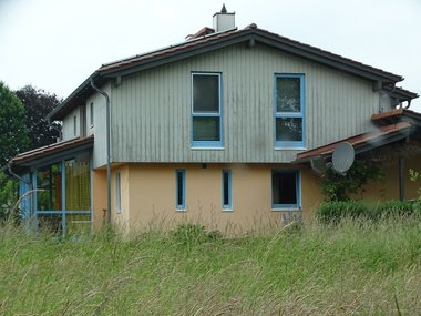 Ein Wohnhaus mit verglaster Veranda in gelb gestrichen mit grünen Holzlatten am Dachstuhl und blauen Fensterrahmen.