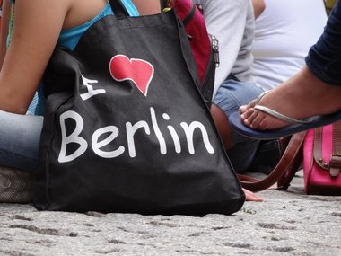 Ein Mädchen in Jens und türkisem schulterfreien T-Shirt sitzt bei einer Vorführung im Schneidersitzt auf hellgrauem Kopfsteinplaster. Über ihrer Schulter hängt eine schwarze Tasche mit weißer Aufschrift "I - rotes Herz - Berlin".