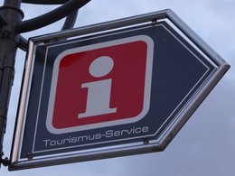 Ein Schild mit einem großen I auf rotem Grund mit einem blauen Rahmen und dem Text Tourismus-Service zeigt nach rechts.