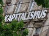 Weisser Schriftzug mit Kapitalismus in riesen Buchstaben an einer alten, verfallenen Häuserwand in Ost-Berlin mit grünen Kastanienästen im Vordergrund.. 