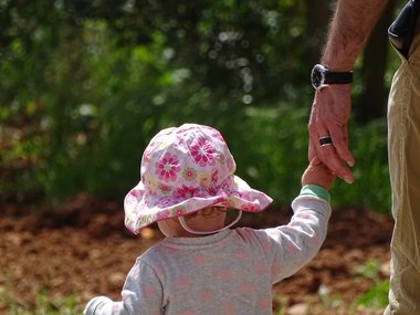 Ein Kleinkind mit einem Sonnenhut mit Blumen in rosa geht an einer Hand mit Uhr und Ring spazieren.