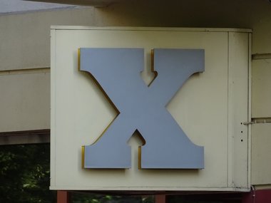 Ein großes plastisches X in grau an einer cremfarbenen Wand.