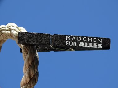 Eine schwarze Wäscheklammer mit der weißen Aufschrift: Mädchen für Alles hängt an einem Seil vor blauem Himmel.