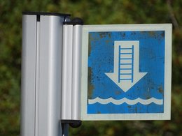 Ein kleines blaues Schild mit einem Pfeil nach unten, wobei auf dem Pfeil eine Leiter und darunter Wassser dargestellt ist.Das Schild ist leicht verdreckt.