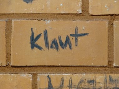 Rote Ziegelsteinmauer in einer Turnhallenkabine auf dem das Wort klaut geschrieben steht.