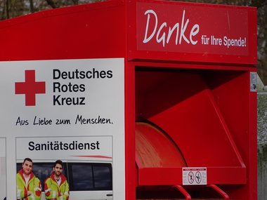 Ein Container vom Roten Kreuz in dem Spenden gesammelt werden und einem seitlich Bild von zwei Männern vom Sanitätsdienst.