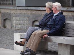 Ein Seniorenpaar mit blauen Jacken sitzt auf einer Parkbank aus Beton.