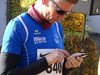 Jogger in blauer Sportkleidung mit Startnummer auf der Brust und Sonnenbrille, der gerade auf seinem Smartphone tippt.