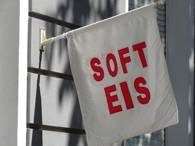 Eine weiße Fahne, die an einer Hauswand befestigt ist, wirbt mit roter Schrift für Softeis.