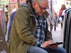 Das Bild zeigt einen älteren Mann mit einem Tablet-PC, der auf etwas sitzt und mit der Hand über das Tablet streicht.