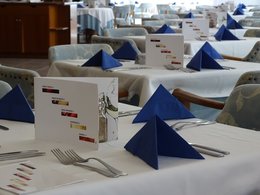 Mehrere gedeckte Tische in einem Restaurant mit blauen Servietten und einer Weinkarte in der Mitte.