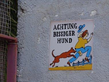 Ein Schild an einer weißen Mauer mit einer Person, die von einem Hund in den Po gebissen wird und der Aufschrift: Achtung bissiger Hund.