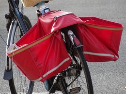 Auf dem Gepäckträger eines abgestellten Fahrrads hängen zwei rote Taschen mit Zeitungen.