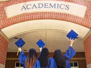 Drei Frauen halten vor ihrer Universität ihren Doktorhut hoch und freuen sich über ihren akademischen Abschluss.